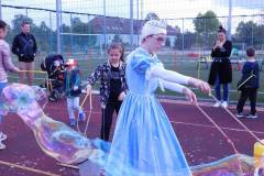 Zdjęcie przedstawia kobietę, która znajduje się na środku zdjęcia i jest przebrana za księżniczkę robi olbrzymie bańki. W oddali jest mężczyzna z wózkiem, a całkowicie po prawej stronie znajduje się kobieta, a przed nią dziewczyna odwrócona plecami.