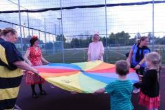 Na zdjęciu została przedstawiona wielka rozłożona kolorowa chusta na boisku od koszykówki. Wokół niej znajdują się dzieci, pani w stroju Myszki Miki, pani w stroju pszczółki Mai oraz pani w różowym stroju z różkami.